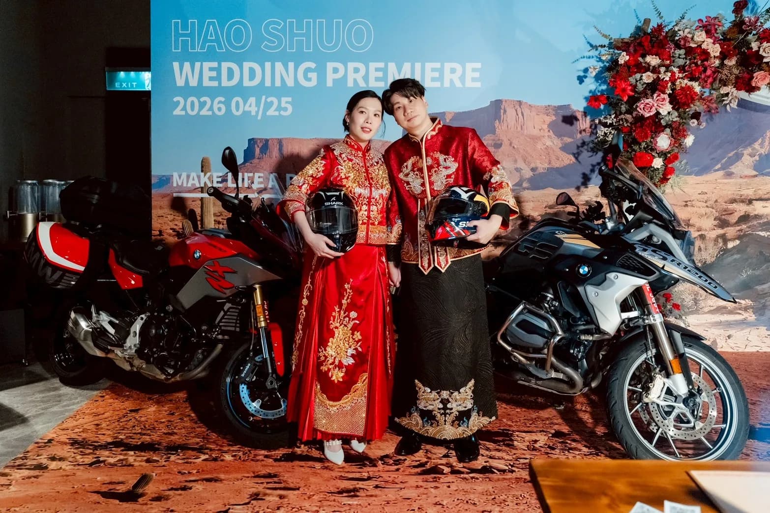 身著紅金中式禮服的新婚夫婦,手持頭盔與摩托車在沙漠背景前合影。